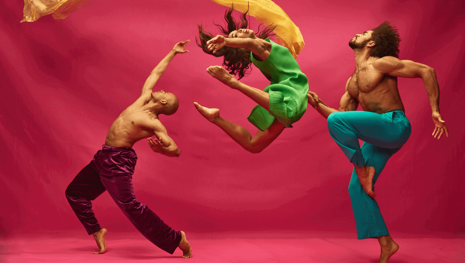 Three dancers in motion wearing colorful clothing against a pinkish red background, the center dancer leaping in the air.